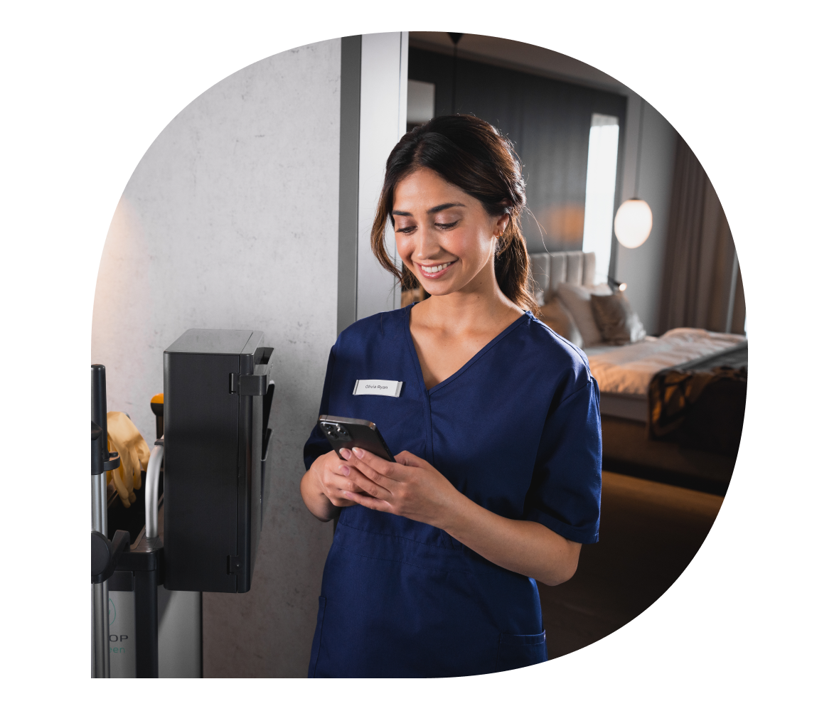 Smiling housekeeping staff member in a navy uniform using a smartphone, standing next to a cleaning cart, with a well-lit hotel room in the background.