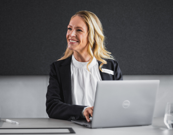 Smiling woman in professional attire sitting at a desk with a laptop, looking to the side