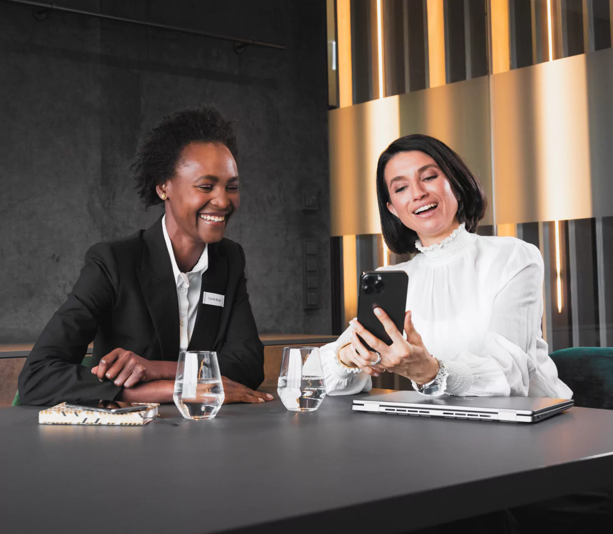 Two professional women sitting at a table in a modern office, laughing and looking at a smartphone. One woman is dressed in a black blazer, and the other in a white blouse, with a laptop, notebook, and glasses of water on the table.