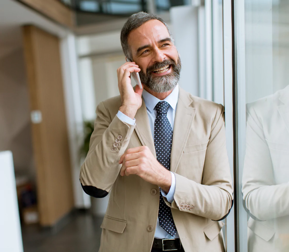 Smiling hotelmanager with a gray beard in a beige suit, talking on a smartphone while standing next to a glass wall in a modern office setting.